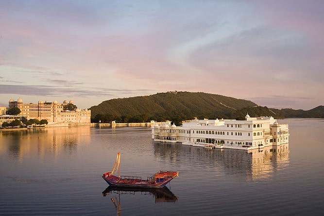 "Lake Pichola in Udaipur, Rajasthan, featuring the iconic Taj Lake Palace and City Palace at sunset with a traditional boat on calm waters."